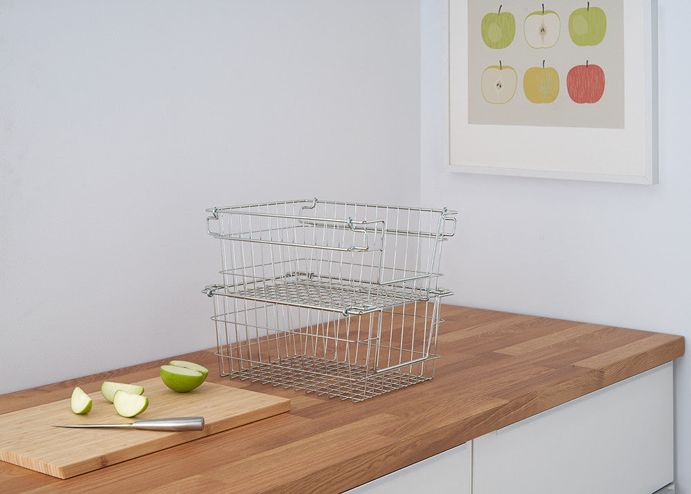 A pair of silver wire baskets are stacked on a wooden countertop. A cutting board with sliced green apples and a knife are visible to the left, and a framed apple print hangs on the white wall behind.