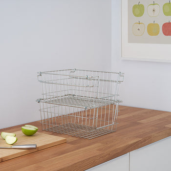 A pair of silver wire baskets are stacked on a wooden countertop. A cutting board with sliced green apples and a knife are visible to the left, and a framed apple print hangs on the white wall behind.
