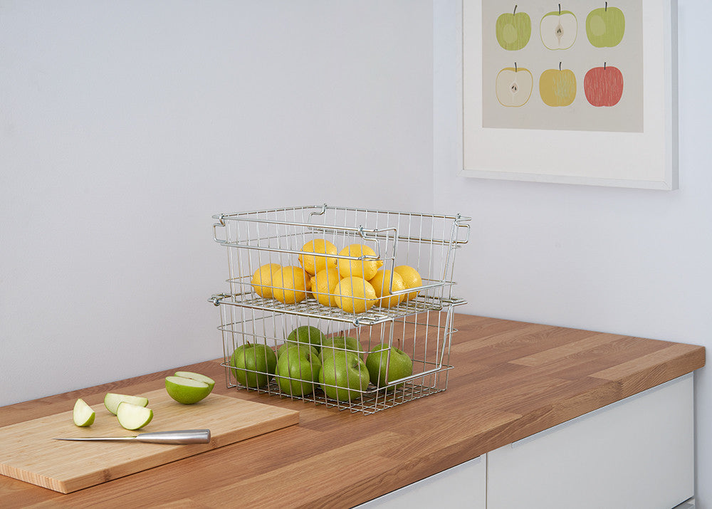 A kitchen counter features a wooden cutting board with sliced green apples and a knife, and two stacked wire baskets filled with lemons and green apples. A framed print of colorful apple illustrations hangs on the white wall above.