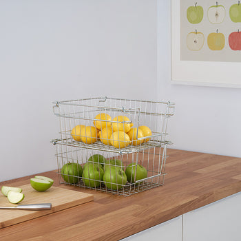 A kitchen counter features a wooden cutting board with sliced green apples and a knife, and two stacked wire baskets filled with lemons and green apples. A framed print of colorful apple illustrations hangs on the white wall above.
