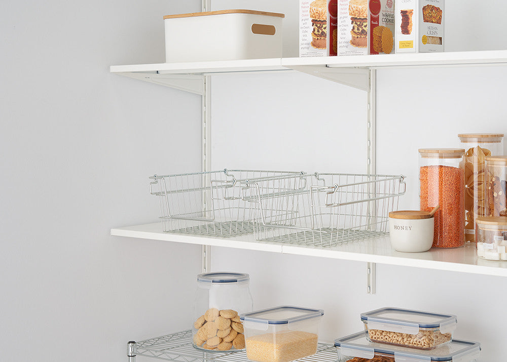 Two empty silver wire baskets sit side-by-side on a white shelf, part of an organized pantry with various food containers. The baskets appear stackable, with handles visible on the sides.