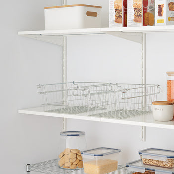 Two empty silver wire baskets sit side-by-side on a white shelf, part of an organized pantry with various food containers. The baskets appear stackable, with handles visible on the sides.
