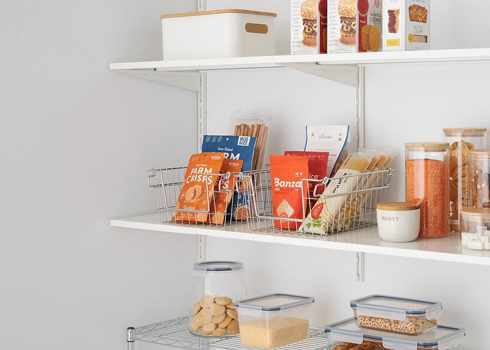 Two wire baskets filled with snacks sit on a white shelf, part of an organized pantry. The shelves also hold clear containers with food, a white box, and various packaged goods.