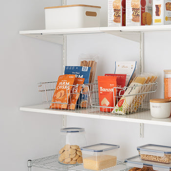 Two wire baskets filled with snacks sit on a white shelf, part of an organized pantry. The shelves also hold clear containers with food, a white box, and various packaged goods.