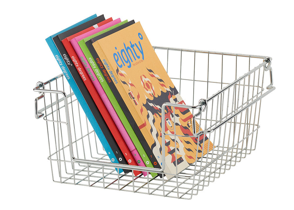 A silver wire basket filled with several colorful magazines titled "eighty degrees" is angled on a white background. The basket's open wire design is visible, and the magazines are neatly stacked inside, showing a variety of cover colors.
