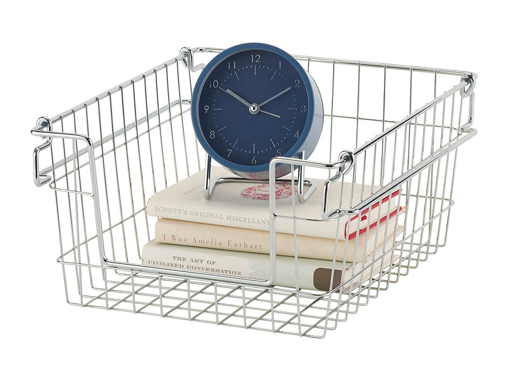 A chrome wire storage basket holds a stack of three books and a blue analog clock with a stand. The basket has a grid pattern base and a smooth, reflective finish.