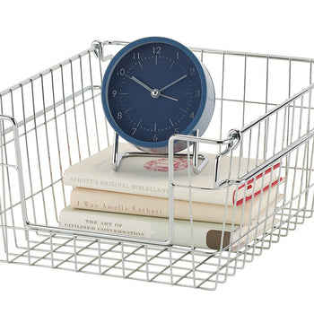 A chrome wire storage basket holds a stack of three books and a blue analog clock with a stand. The basket has a grid pattern base and a smooth, reflective finish.