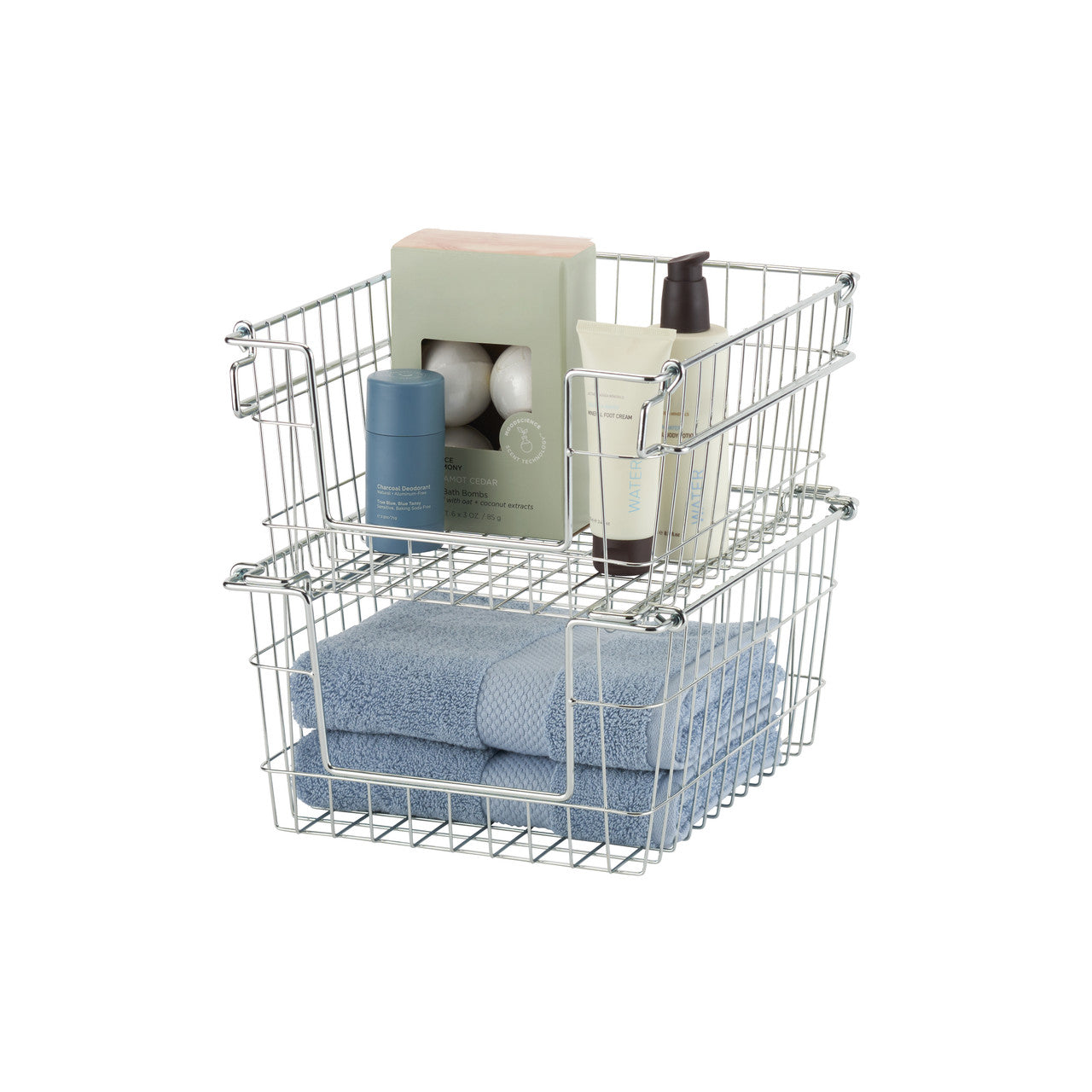 A photo of two stacked, rectangular wire baskets with chrome finish, containing bathroom items. The top basket holds a box of bath bombs, a tube of lotion, and a deodorant stick, while the bottom basket holds folded blue towels.