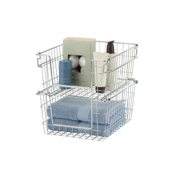 A photo of two stacked, rectangular wire baskets with chrome finish, containing bathroom items. The top basket holds a box of bath bombs, a tube of lotion, and a deodorant stick, while the bottom basket holds folded blue towels.