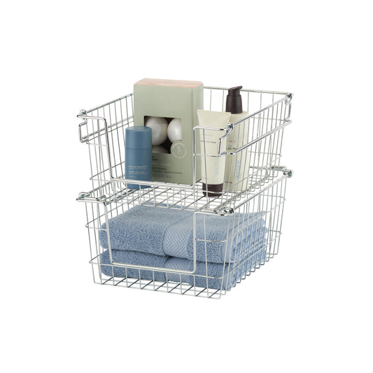 A photo of two stacked, rectangular wire baskets with chrome finish, containing bathroom items. The top basket holds a box of bath bombs, a tube of lotion, and a deodorant stick, while the bottom basket holds folded blue towels.