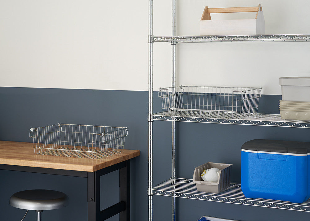 A well-organized workspace shows two wire storage baskets, one on a wooden table and the other on a metal wire shelf, amidst a neutral-toned room with a blue accent wall. The baskets are open, allowing for easy viewing and access to stored items.