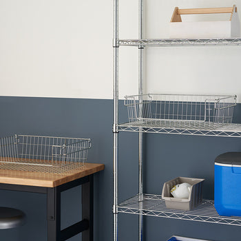 A well-organized workspace shows two wire storage baskets, one on a wooden table and the other on a metal wire shelf, amidst a neutral-toned room with a blue accent wall. The baskets are open, allowing for easy viewing and access to stored items.