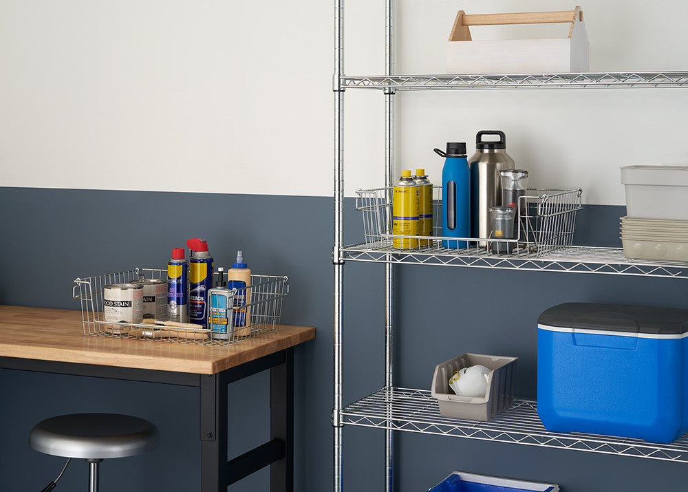 A medium shot of a well-organized garage or workshop space featuring a 5-tier wire shelving rack on the right. The rack holds various items, including spray cans, bottles, and boxes, while a wooden workbench with a wire basket filled with supplies sits on the left.
