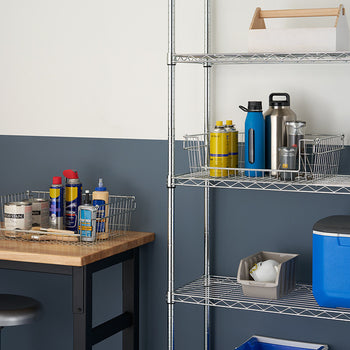 A medium shot of a well-organized garage or workshop space featuring a 5-tier wire shelving rack on the right. The rack holds various items, including spray cans, bottles, and boxes, while a wooden workbench with a wire basket filled with supplies sits on the left.