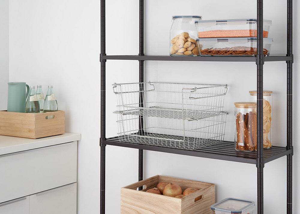 A close-up shot of two stackable, silver metal wire baskets on a black metal shelving unit, used for kitchen organization. The baskets are empty and nested, with various filled glass and plastic containers on the shelves above and beside them.