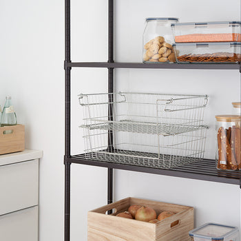 A close-up shot of two stackable, silver metal wire baskets on a black metal shelving unit, used for kitchen organization. The baskets are empty and nested, with various filled glass and plastic containers on the shelves above and beside them.