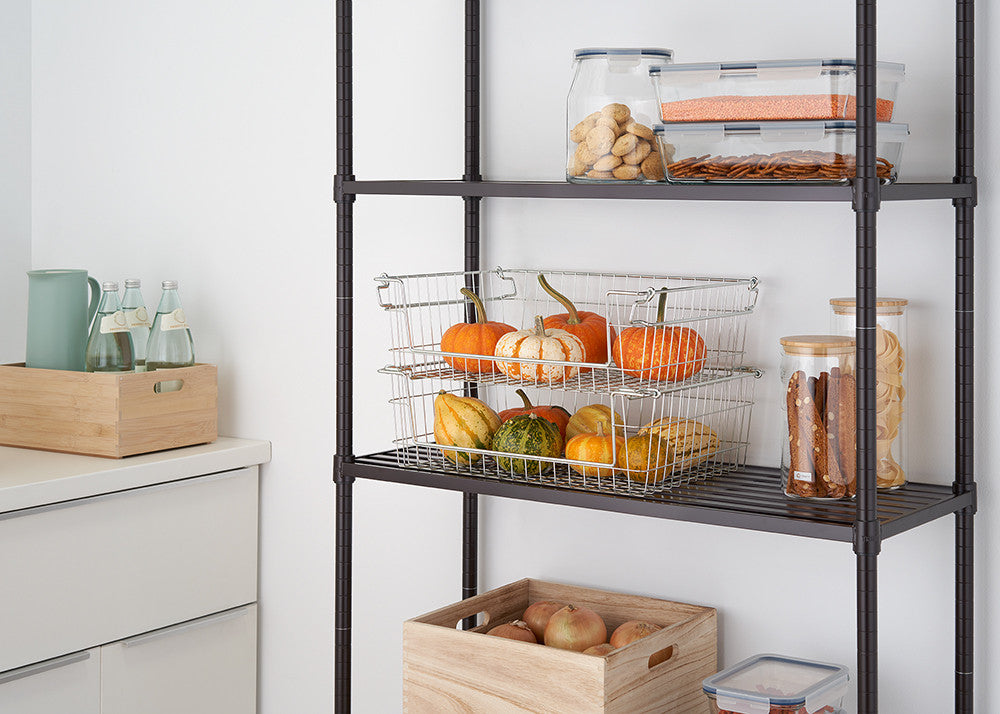 A close-up shot shows two stacked wire baskets filled with small pumpkins and gourds on a dark metal shelving unit. Clear glass containers filled with snacks are visible on the shelf above, and a wooden box holding apples sits on the floor below.