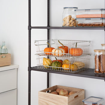 A close-up shot shows two stacked wire baskets filled with small pumpkins and gourds on a dark metal shelving unit. Clear glass containers filled with snacks are visible on the shelf above, and a wooden box holding apples sits on the floor below.