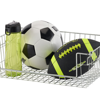 A chrome wire basket contains a green water bottle, a soccer ball, and a football. The basket is rectangular with a grid pattern and has handles on the sides.