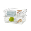 A studio shot of two chrome wire storage baskets stacked on top of each other, filled with various bathroom items. The baskets feature a grid pattern and are shown against a white background.
