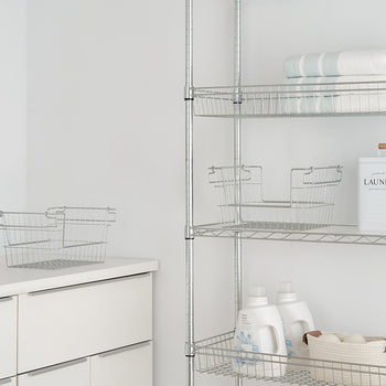 A well-organized laundry room features a metal shelving unit stocked with neatly folded towels, detergent bottles, a laundry container, and wire baskets. A white cabinet with drawers sits below a wall sign displaying 