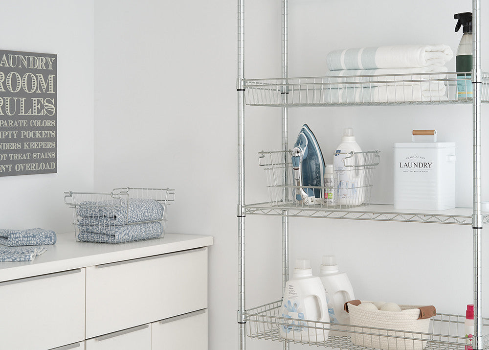 A well-organized laundry room features a white cabinet with folded blue towels in wire baskets and a metal shelving unit storing laundry essentials, including detergent bottles, an iron, and neatly folded linens. A decorative "Laundry Room Rules" sign hangs on the wall, adding a stylish touch to the clean and minimalistic space.