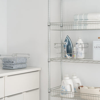A well-organized laundry room features a white cabinet with folded blue towels in wire baskets and a metal shelving unit storing laundry essentials, including detergent bottles, an iron, and neatly folded linens. A decorative 