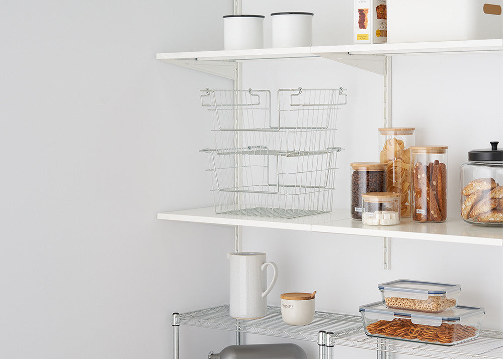 A neatly organized pantry with white shelves holding glass jars with bamboo lids, metal baskets, and storage containers. The jars contain pasta, coffee beans, sugar cubes, and baked goods, while the plastic containers store pretzels and cereal.