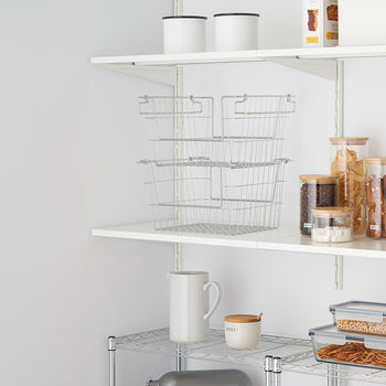 A neatly organized pantry with white shelves holding glass jars with bamboo lids, metal baskets, and storage containers. The jars contain pasta, coffee beans, sugar cubes, and baked goods, while the plastic containers store pretzels and cereal.