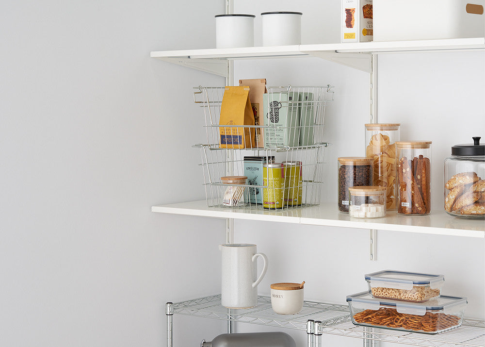 A neatly organized pantry with white shelves holding glass jars with bamboo lids filled with dry food items, metal wire baskets containing packaged goods, and storage containers. A metal rack below the shelves holds a white ceramic mug, a honey jar, and plastic food storage boxes with snacks.