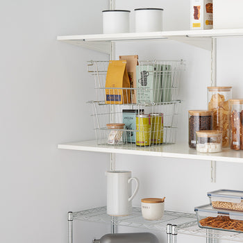A neatly organized pantry with white shelves holding glass jars with bamboo lids filled with dry food items, metal wire baskets containing packaged goods, and storage containers. A metal rack below the shelves holds a white ceramic mug, a honey jar, and plastic food storage boxes with snacks.
