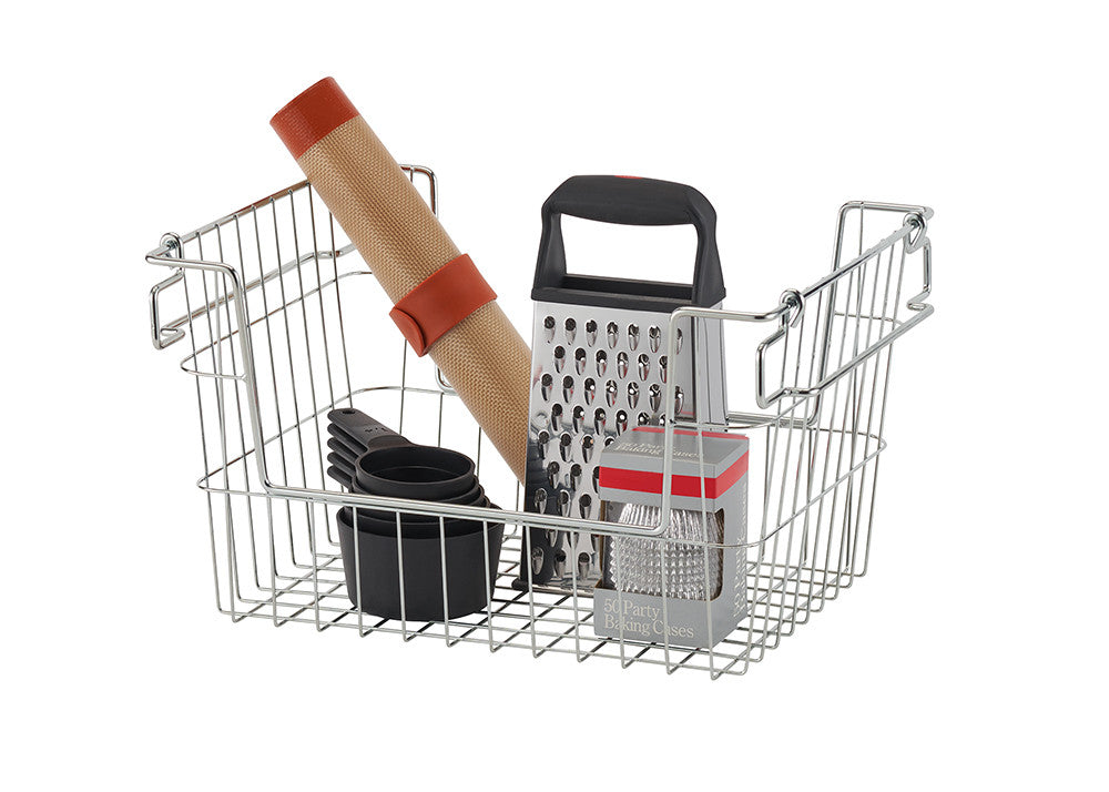 A metal wire basket contains various kitchen tools, including a box grater with a black handle, a rolled-up silicone baking mat secured with a red band, a set of black measuring cups, and a box of silver party baking cases. The basket has collapsible handles and an open design for easy storage.