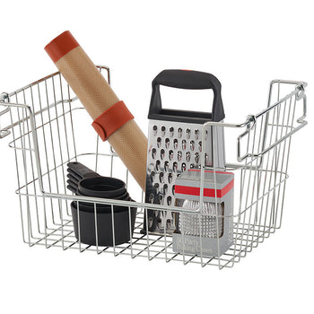 A metal wire basket contains various kitchen tools, including a box grater with a black handle, a rolled-up silicone baking mat secured with a red band, a set of black measuring cups, and a box of silver party baking cases. The basket has collapsible handles and an open design for easy storage.