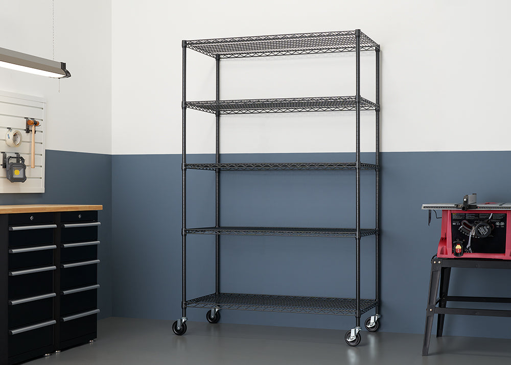 A tall, five-shelf wire shelving unit with locking wheels stands in the middle of a garage workshop. The shelves are empty and the unit is positioned in front of a two-toned wall, with the upper half painted white and the lower half a dark, dusty blue.