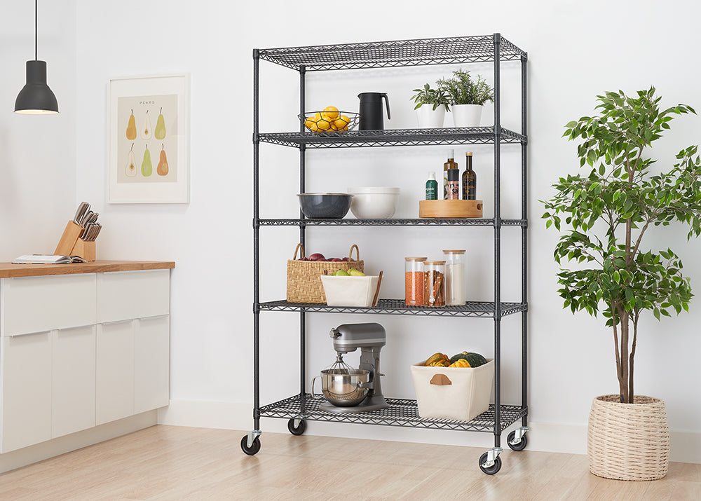 A tall, five-shelf wire rack on wheels stands in a modern kitchen. The shelves are organized with various kitchen items, including bowls, food ingredients, a stand mixer, and decor like a potted plant and a painting.