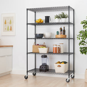 A black metal five-tier wire rolling shelf unit stands in a brightly lit, modern kitchen alongside white cabinetry and a potted houseplant. The open shelves hold a collection of organized essentials, including small potted plants, oil bottles, clear storage jars, a countertop blender, and woven storage baskets.
