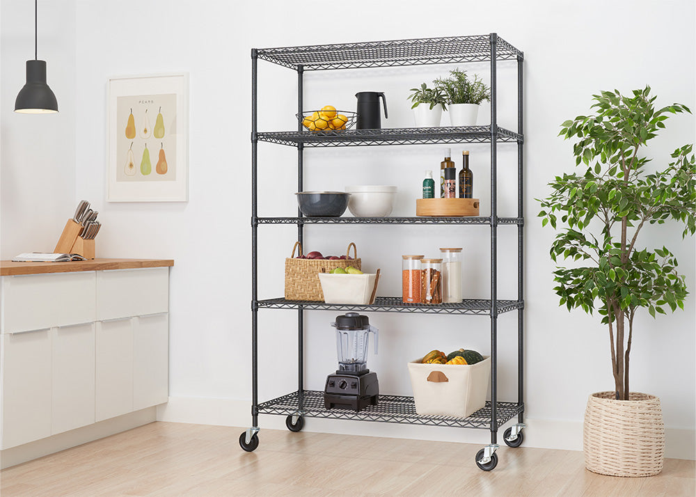 A black metal five-tier wire rolling shelf unit stands in a brightly lit, modern kitchen alongside white cabinetry and a potted houseplant. The open shelves hold a collection of organized essentials, including small potted plants, oil bottles, clear storage jars, a countertop blender, and woven storage baskets.