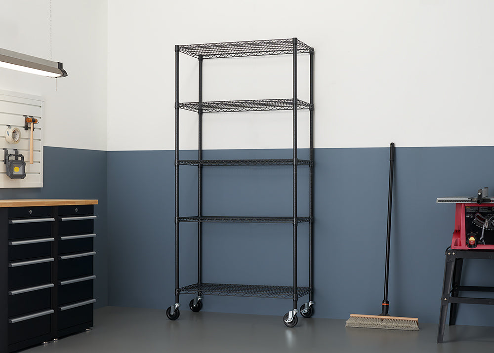 A tall, black, five-tier wire shelving unit with caster wheels stands against a wall painted white over dark blue-gray. To the right of the shelf, a broom rests against the wall.