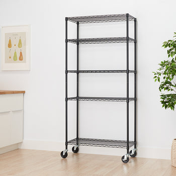 A tall, black wire shelving unit with five shelves is pictured against a white wall and light wood floor. The unit is on casters for mobility and stands next to a potted plant in a woven basket.