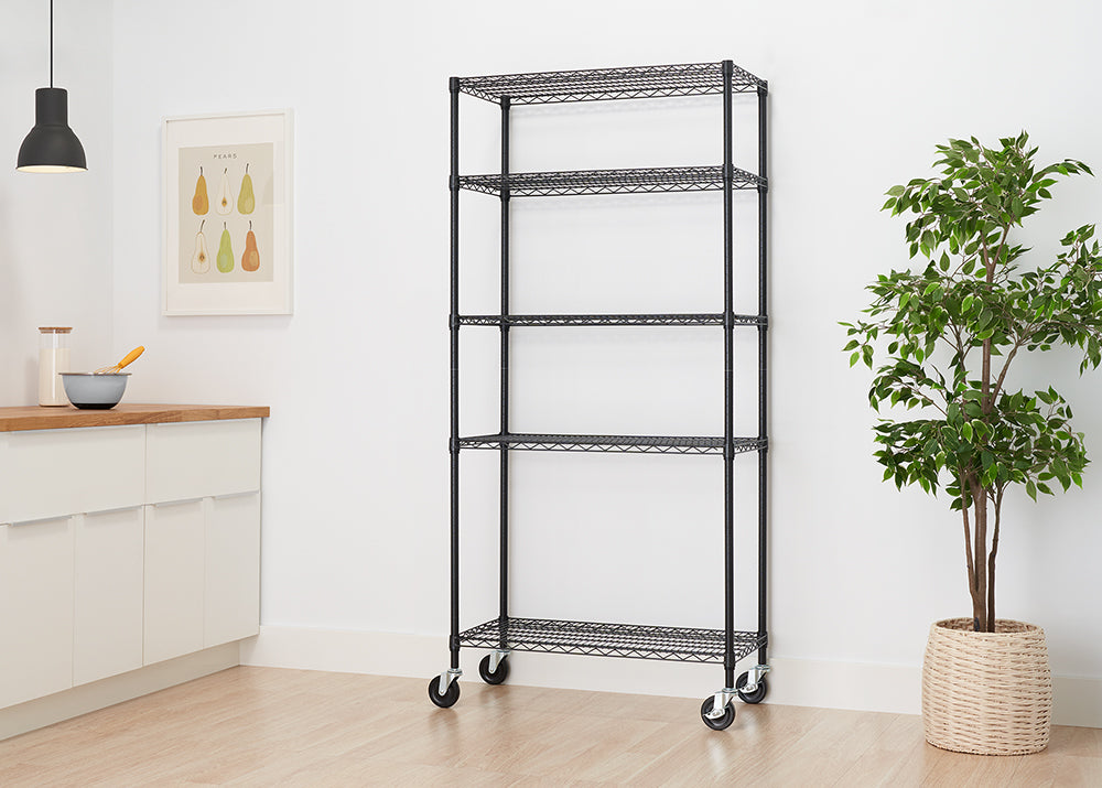 A tall, black wire shelving unit with five shelves is pictured against a white wall and light wood floor. The unit is on casters for mobility and stands next to a potted plant in a woven basket.