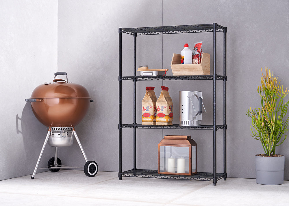 A shiny copper kettle grill with a metal base and two wheels sits next to a black, four-tier wire shelving unit. The shelves hold various items, including a wooden crate with bottles, two bags of charcoal, a chimney starter, and a copper and glass lantern with candles.