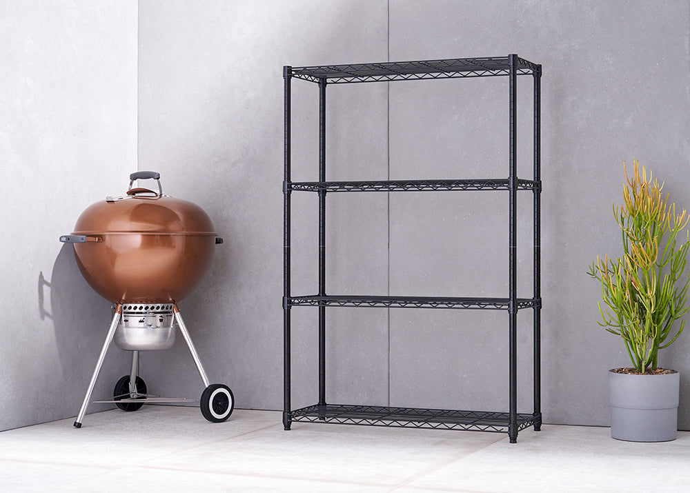 A four-tiered black wire shelving unit stands next to a shiny copper-colored charcoal grill with wheels. A green succulent plant in a gray pot is on the opposite side of the shelving unit.