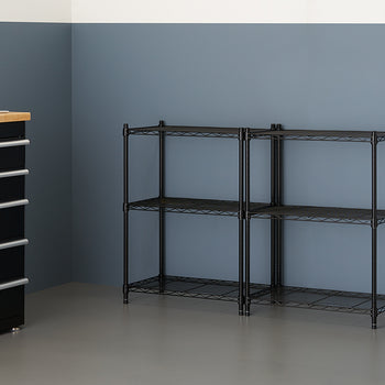 A clean garage or utility room features two black wire shelving units placed side-by-side against a muted blue wall. To the left, a black rolling tool chest with a wooden top and a bottle of yellow liquid sits near the shelving.