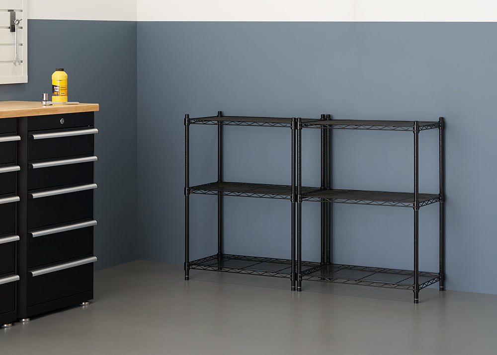 A clean garage or utility room features two black wire shelving units placed side-by-side against a muted blue wall. To the left, a black rolling tool chest with a wooden top and a bottle of yellow liquid sits near the shelving.