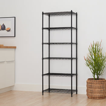 A tall, narrow, black wire shelving unit with six shelves stands against a plain white wall on a light wood floor. A large potted plant in a wicker basket is placed to the right of the shelving unit.