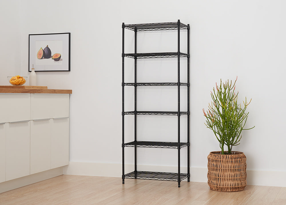 A tall, narrow, black wire shelving unit with six shelves stands against a plain white wall on a light wood floor. A large potted plant in a wicker basket is placed to the right of the shelving unit.