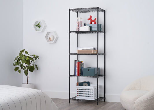 A five-tier black wire shelving unit stands against a white wall, organized with various items including books, storage bins, and a small red dog figurine. The minimalist room also features a potted plant, two hexagonal wall shelves, and the edge of a bed with striped bedding.