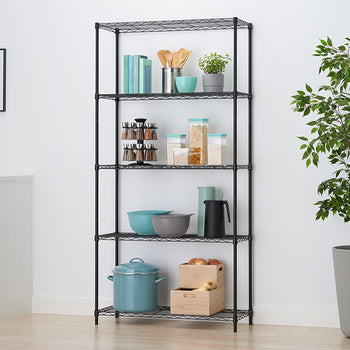 A black wire shelving unit stands organized with various kitchen and pantry items in a brightly lit kitchen setting. Shelves hold items like a rotating spice rack, teal kitchenware, wooden boxes of onions, cookbooks, and dry good containers, positioned next to a tall potted plant and white cabinets.
