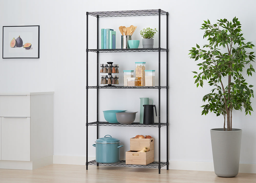 A black wire shelving unit stands organized with various kitchen and pantry items in a brightly lit kitchen setting. Shelves hold items like a rotating spice rack, teal kitchenware, wooden boxes of onions, cookbooks, and dry good containers, positioned next to a tall potted plant and white cabinets.
