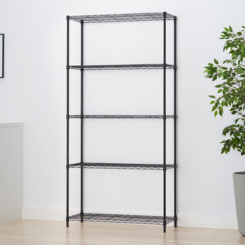 A black wire shelving unit stands empty in a brightly lit room next to a potted Ficus tree and a white sideboard, featuring five adjustable wire shelves. A framed art print of figs hangs on the white wall.